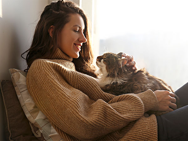A person with long hair, wearing a brown sweater, is sitting by a sunny window. They are smiling and gently caressing a fluffy cat that is sitting on their lap. The cat appears content and is looking up at the person, who could easily be mistaken for a caring veterinarian. Pillows are visible beside them.