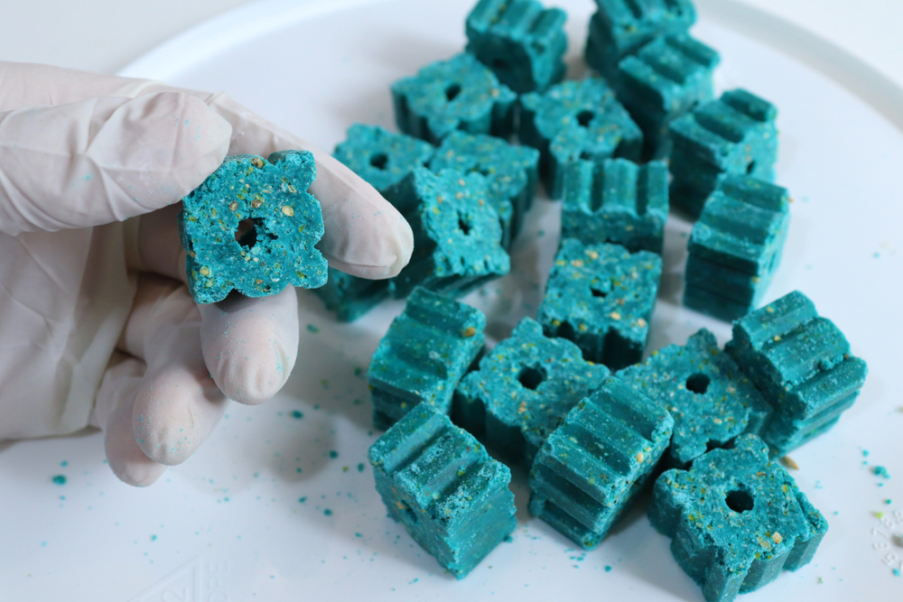 A gloved hand, reminiscent of those worn by a veterinarian during an examination, holds up a blue block with a hole in the center. More identical blue blocks are scattered on a white surface in the background. The blocks have a rough texture and appear to be made of a compressed material.