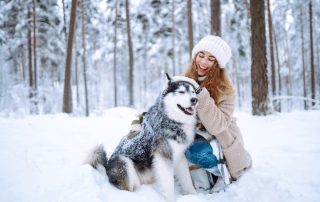 A woman wearing a white knitted hat and beige coat kneels in the snow next to a large, fluffy Siberian Husky. They are in a snow-covered forest, both smiling as she pets the dog lovingly. Snow clings to the dog's fur, showing their close bond—she's not just an owner but a caring veterinarian.