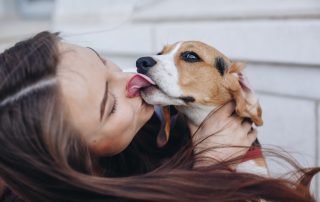 A woman with long brown hair is holding a small dog with brown and white fur. The dog, recently treated by the vet, is licking her face, and the woman is smiling with her eyes closed. They appear to be sitting outside in front of a white building.