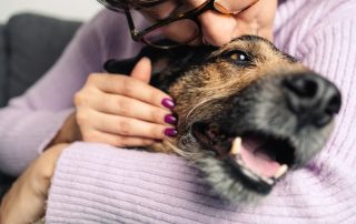 A veterinarian in glasses hugs a smiling dog closely, pressing their cheek against the dog's head. The person has painted fingernails and is wearing a lavender sweater. The dog appears relaxed and happy. The background is slightly blurred.