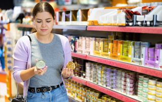 A woman with dark hair, wearing a grey shirt and a light purple cardigan, holds two cans while shopping in a pet food aisle. She appears to be comparing the items, possibly considering advice from her veterinarian. Shelves filled with various pet food products are visible in the background.