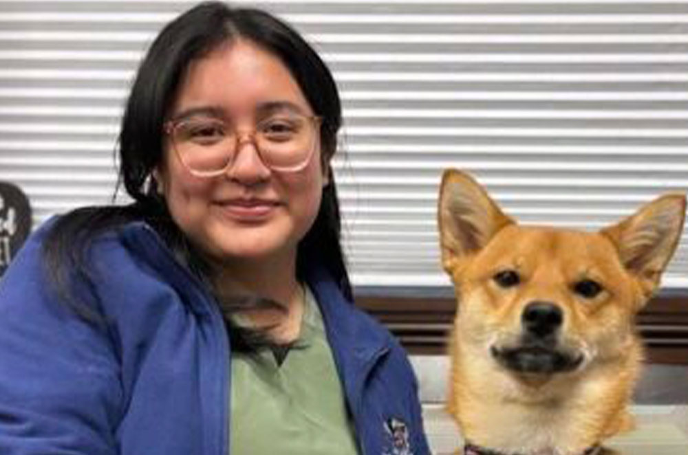 A woman wearing glasses and a blue jacket smiles while seated next to a brown dog with pointy ears. The background features horizontal blinds, giving the impression of a relaxed moment at the vet's office.