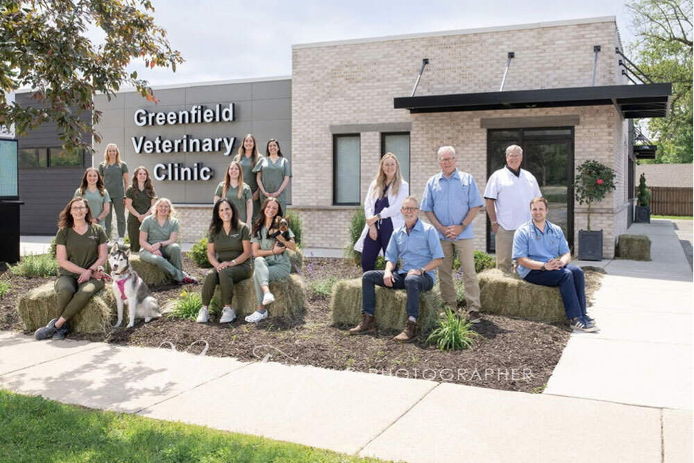 A group of people, some seated and some standing, pose outside the Greenfield Veterinary Clinic building on a sunny day. A dog sits with them. Everyone is smiling and dressed in casual or scrubs attire.